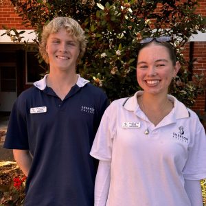 A male and female student stand smiling for the camera in front of a leafy background.