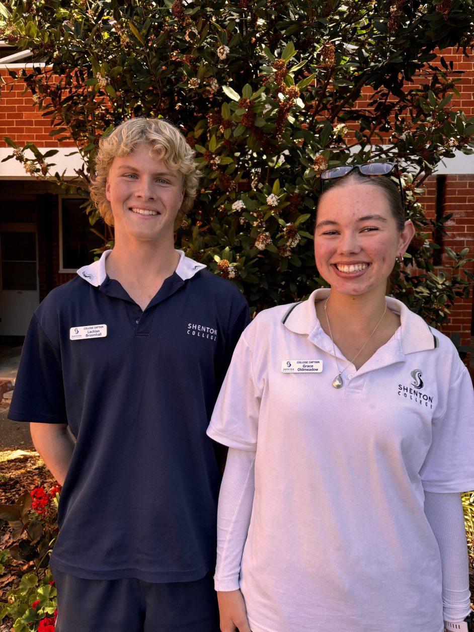 A male and female student stand smiling for the camera in front of a leafy background.